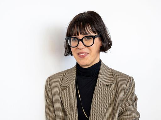Eva Karl Faltermeier steht mit schwarzem Oberteil und kariertem Sakko vor einem weißen Hintergrund. Sie guckt durch eine schwarz umrandete Brille in die Kamera.