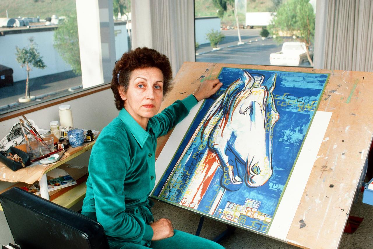 Françoise Gilot sitzt neben einem Gemälde mit der Darstellung eines blauen Pferdes am Tisch.