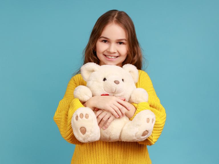 Ein Mädchen mit langen Haaren und im gelben Pullover umarmt einen Teddybär