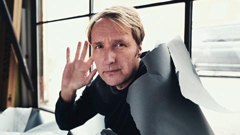 Jens Thomas scheint aus einem Karton zu brechen und hebt die Hand, als ob er intensiv lauschen würde.