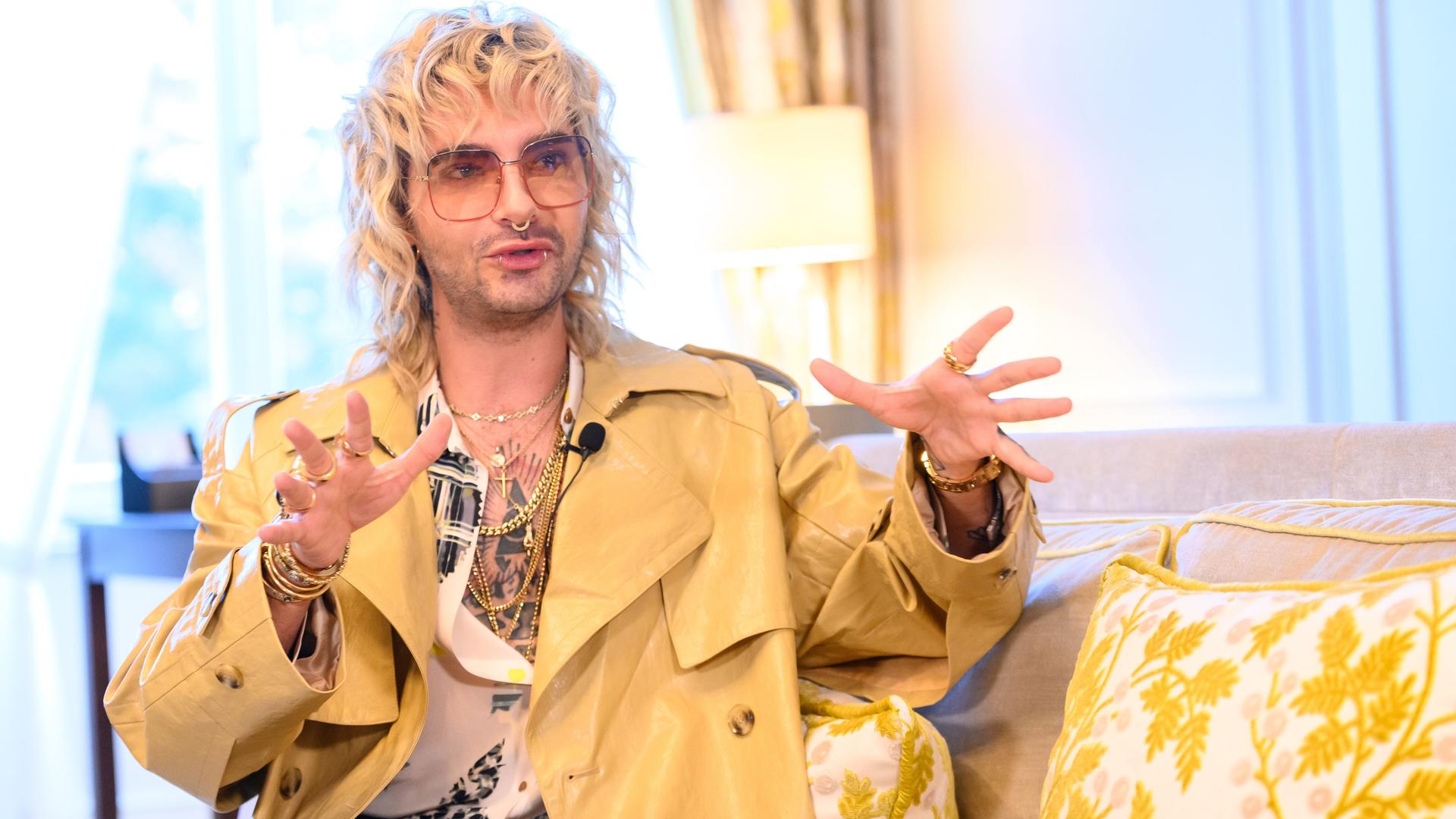Tokio-Hotel-Sänger Bill Kaulitz sitzt in einer Suite des Hotel Vier Jahreszeiten in Hamburg auf einem Sessel und schaut in die Kamera. Er hat blonde, lockige Haare, trägt eine Brille und einen gelblichen Mantel, das Hemd ist weit offen.