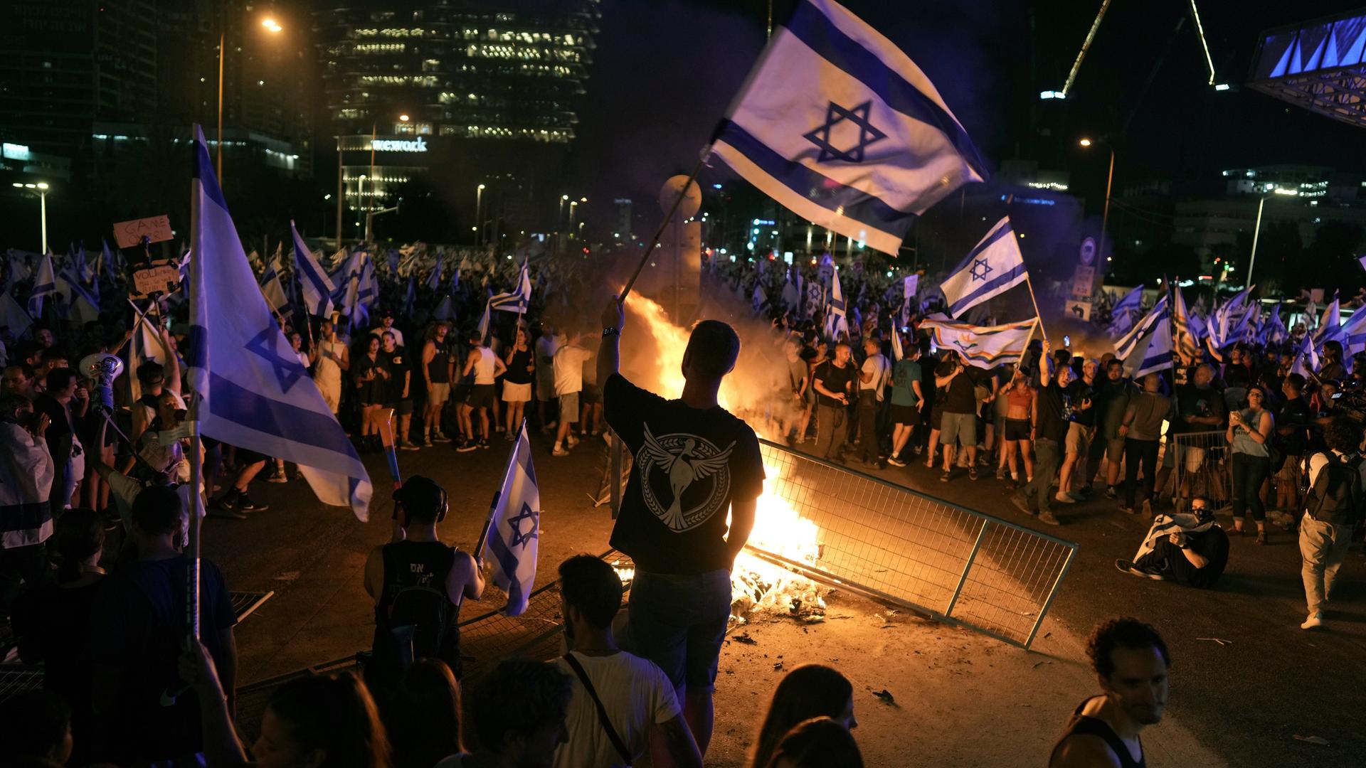 Zahlreiche Menschen mit Israel-Flaggen stehen bei Nacht um ein Feuer auf einer Straße. In dem Feuer liegt eine Metallabsperrung. Im Vordergrund sieht man die Silhouette eines jungen Mannes von hinten. Er schwenkt eine Israel-Flagge.