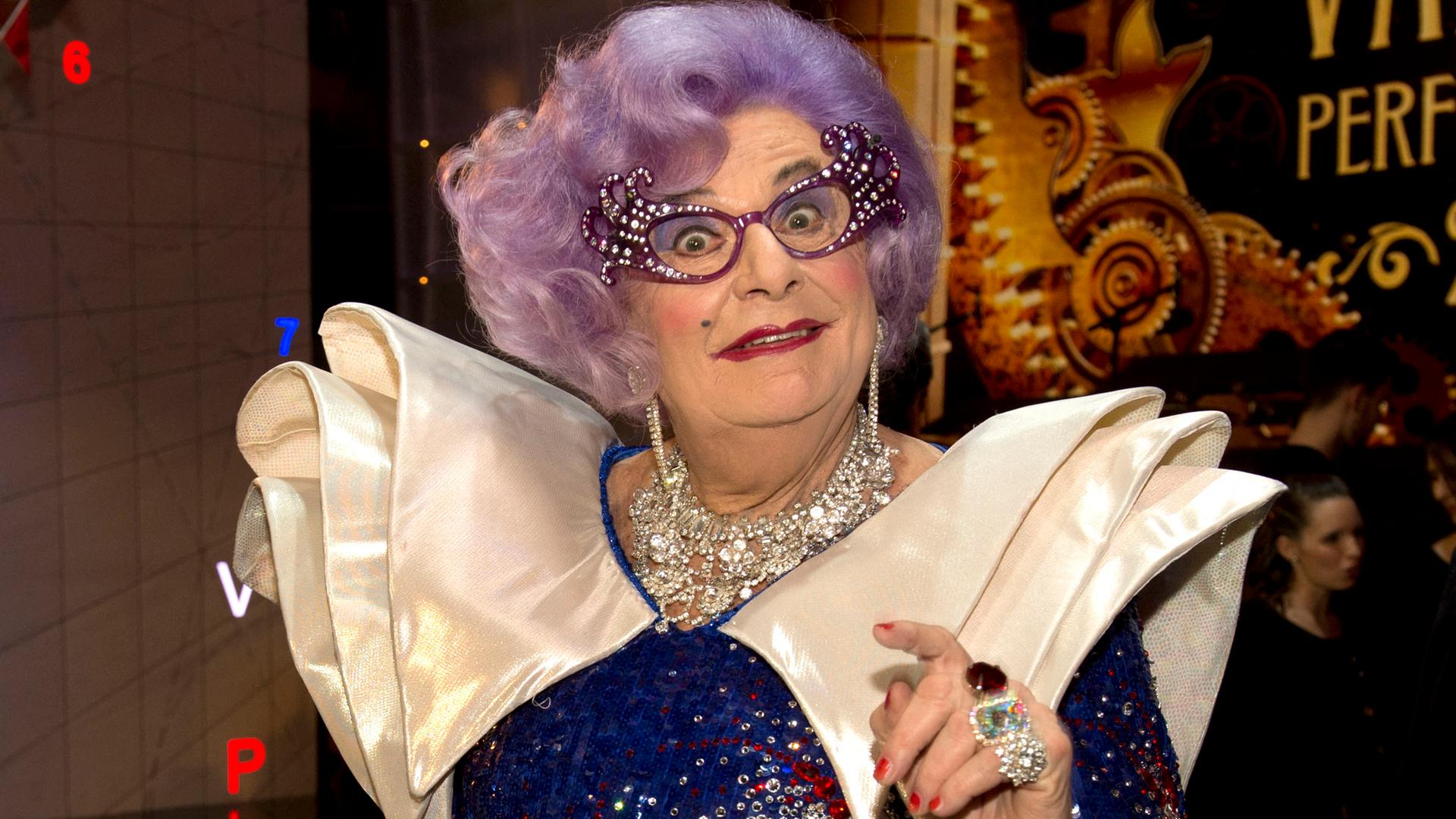Das ist der Komiker Barry Humphries in seiner Rolle als Dame Edna Everage. Er trägt eine lila Perücke und eine strassbesetzte Brille.
