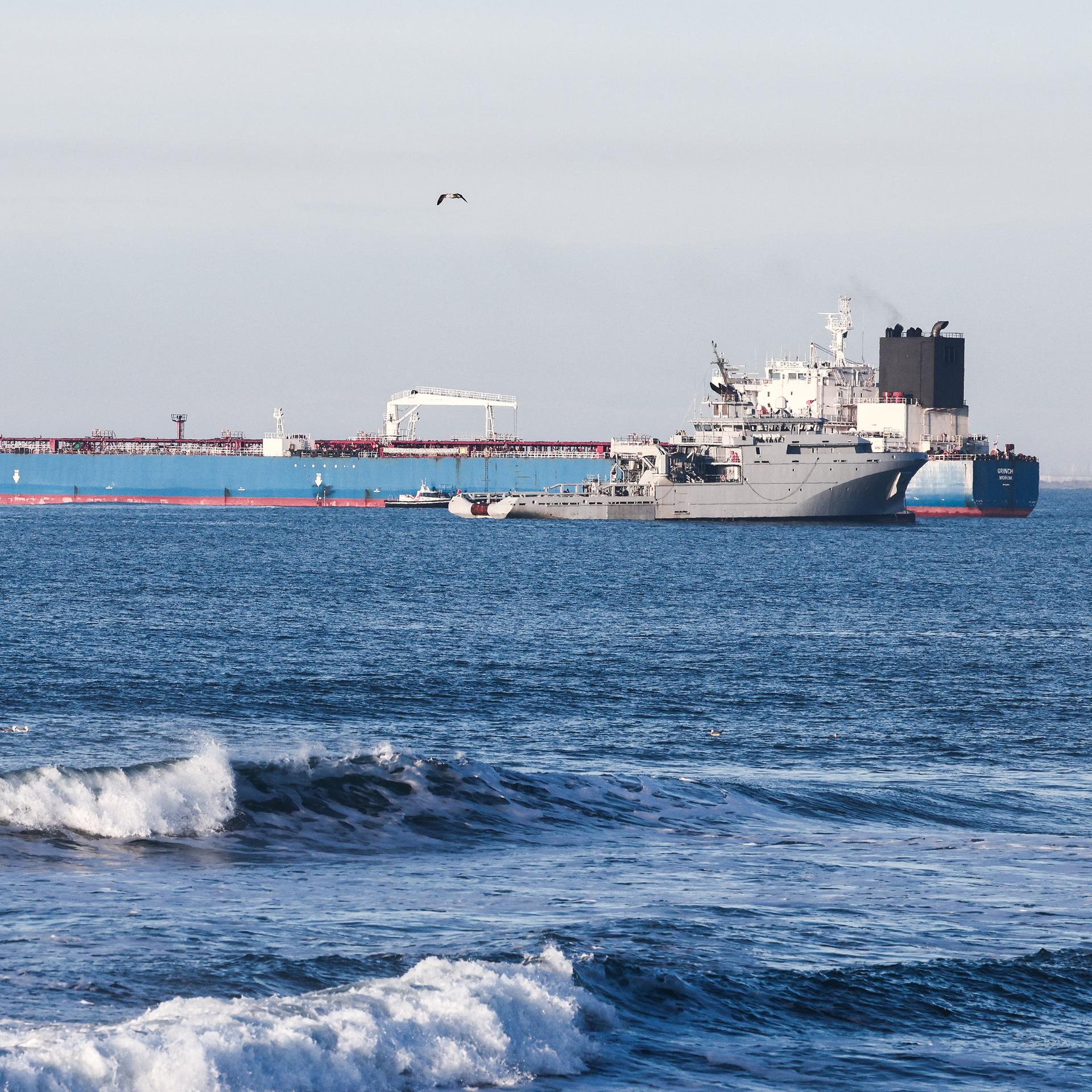 Blick auf einen Öltanker mit blauem Rumpf. Davor kreuzt ein französisches Kriegsschiff