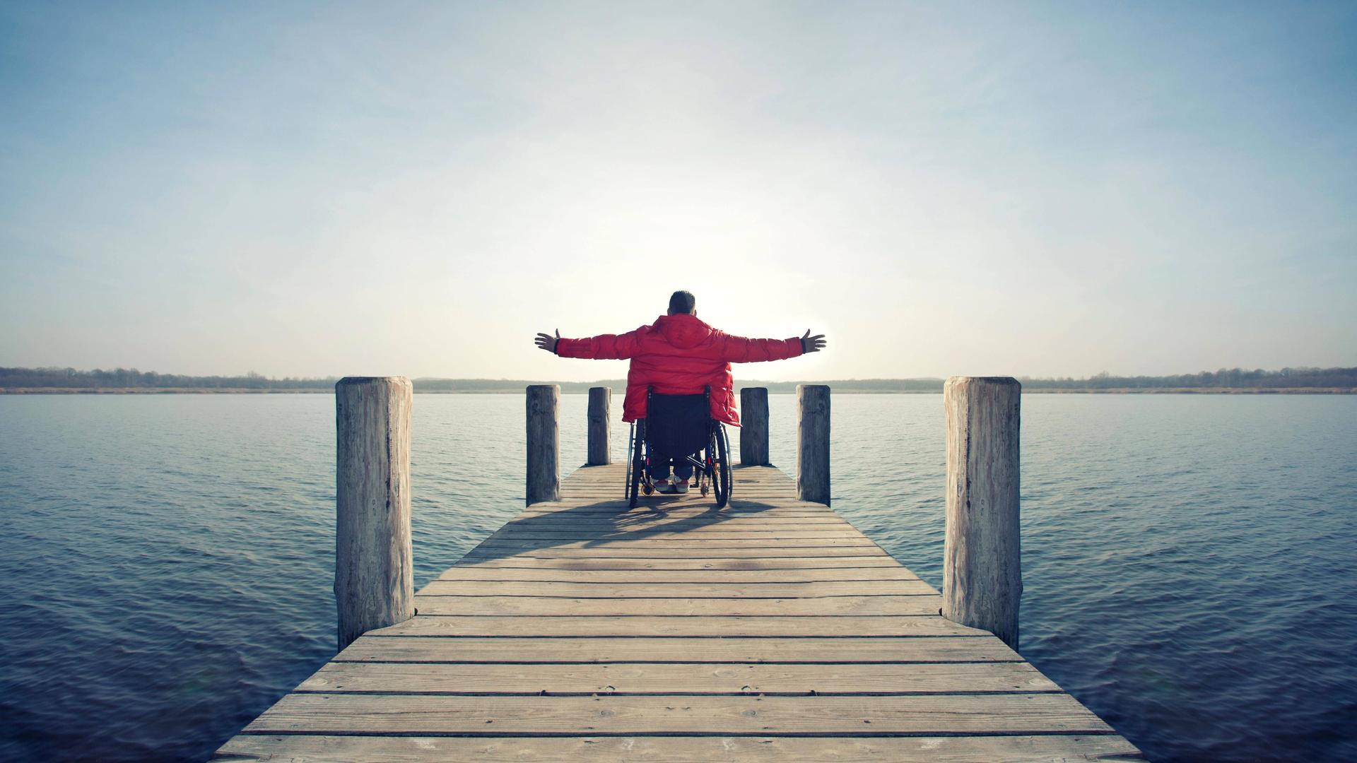 Ein Rollstuhlfahrer auf einem Bootssteg an einem See breitet die Arme aus.