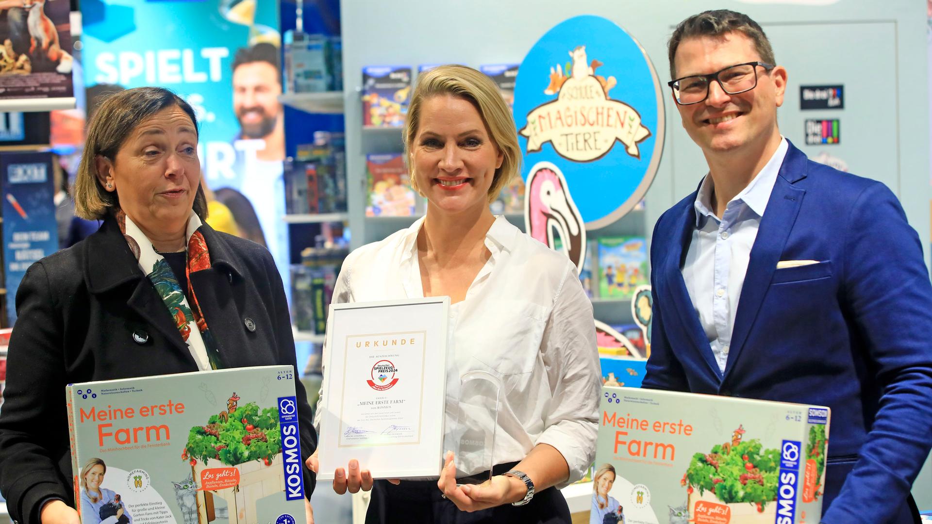 Judith Rakers steht auf der Frankfurter Buchmesse mit einem Preis in einem Bilderrahmen vor einem Bücherstand neben zwei weiteren Personen.