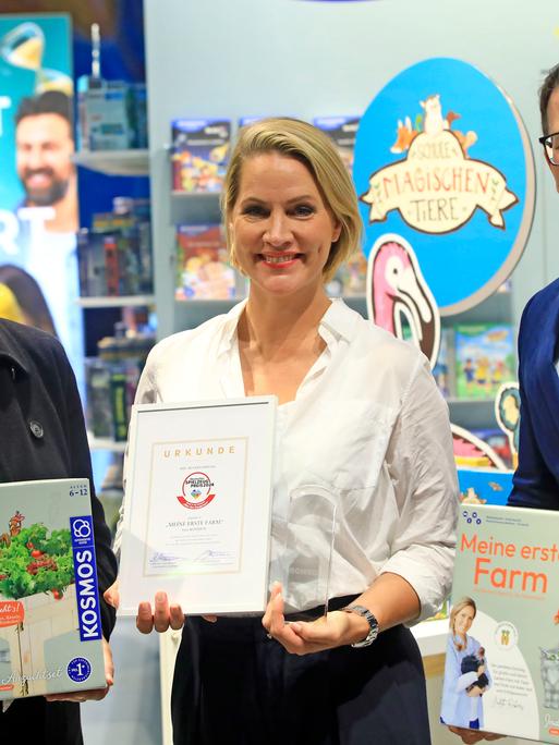 Judith Rakers steht auf der Frankfurter Buchmesse mit einem Preis in einem Bilderrahmen vor einem Bücherstand neben zwei weiteren Personen.