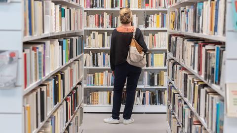 Eine Frau steht vor Bücherregalen in einer Bibliothek und betrachtet die Buchrücken. 