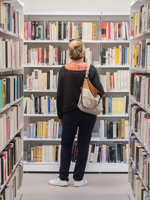 Eine Frau steht vor Bücherregalen in einer Bibliothek und betrachtet die Buchrücken. 