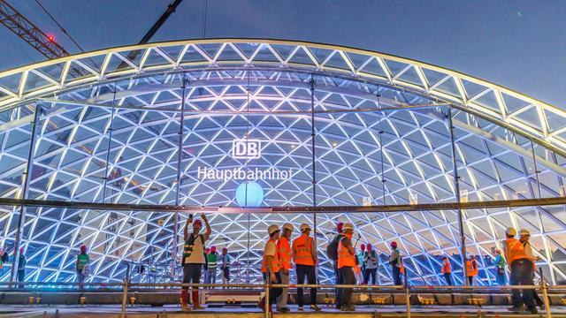 Das Foto zeigt einen Teil des neuen Bahnhofs. Davor stehen einige Menschen. Sie tragen Warnwesten.