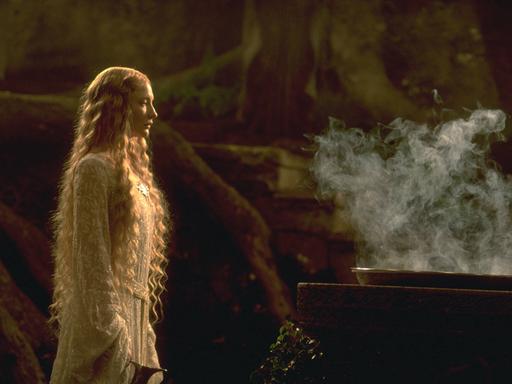 Cate Blanchett als Galadriel steht in einem weißen Kleid vor einem rauchenden Kessel.