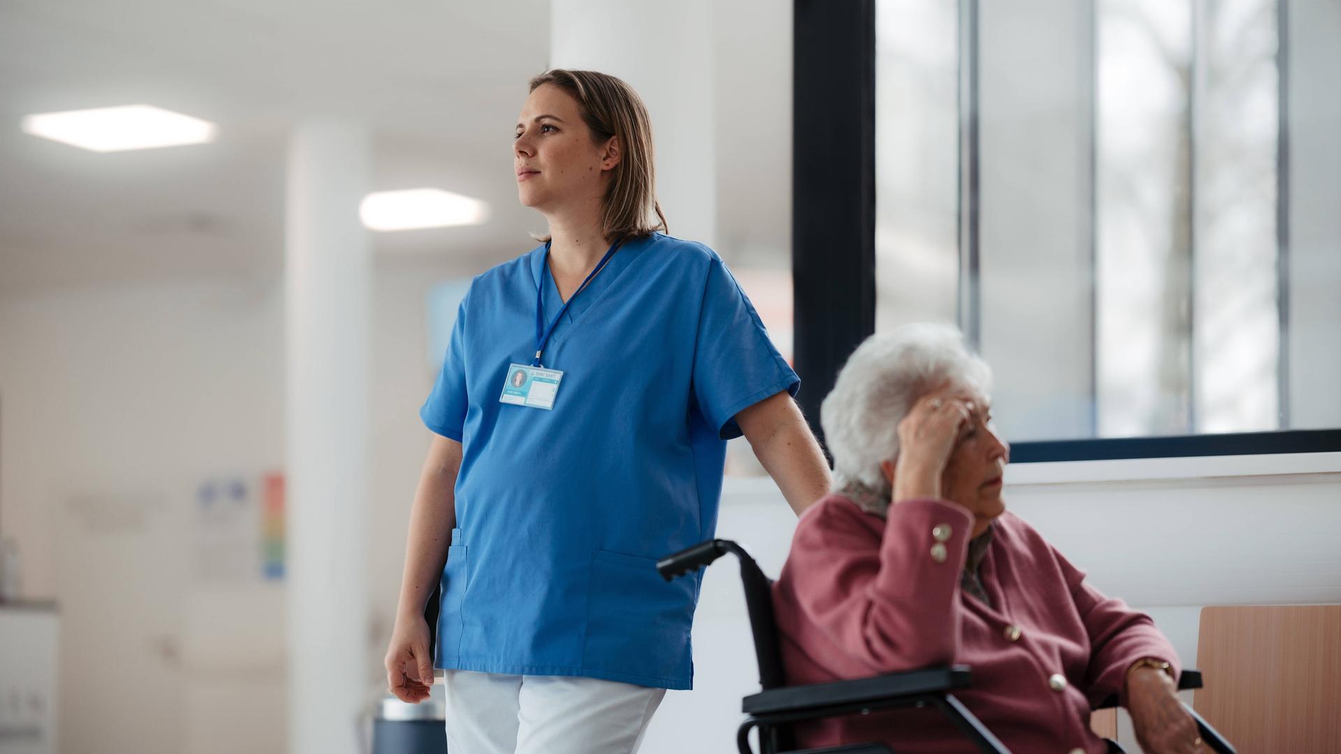 Eine müdeKrankenschwester wartet mit einer älteren Patientin im Rollstuhl im Krankenhausflur. 