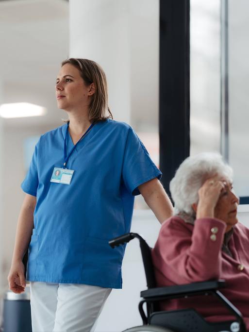 Eine müdeKrankenschwester wartet mit einer älteren Patientin im Rollstuhl im Krankenhausflur. 