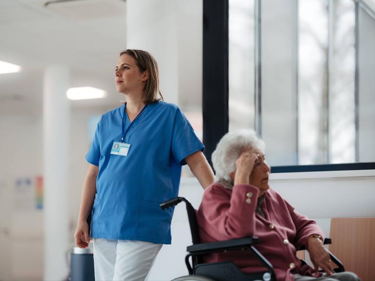Eine müdeKrankenschwester wartet mit einer älteren Patientin im Rollstuhl im Krankenhausflur. 