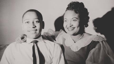 Schwarzweiß-Familienschnappschuss: Emmett Till sitzt fröhlich neben seiner Mutter Mamie Bradley, die freundlich lacht.