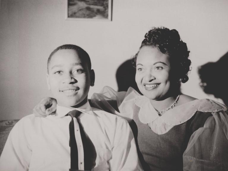 Schwarzweiß-Familienschnappschuss: Emmett Till sitzt fröhlich neben seiner Mutter Mamie Bradley, die freundlich lacht.