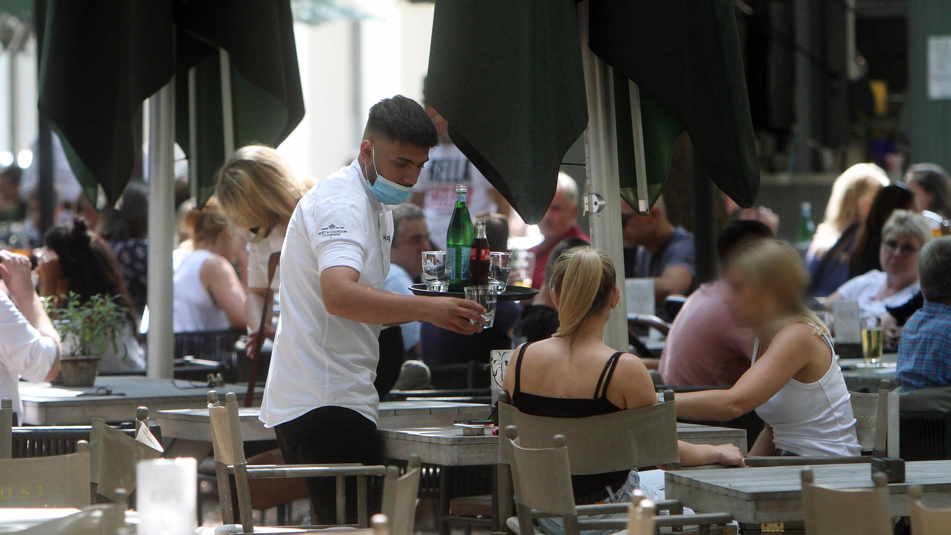 Ein Kellner mit Getränken auf einem Tablett bedient Gäste an einem Tisch im Außenbereich eines Restaurants.