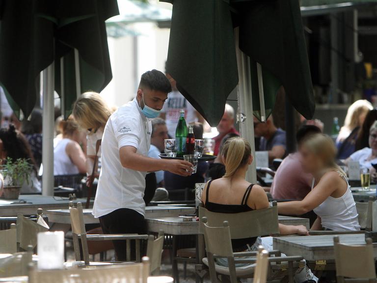Ein Kellner mit Getränken auf einem Tablett bedient Gäste an einem Tisch im Außenbereich eines Restaurants.