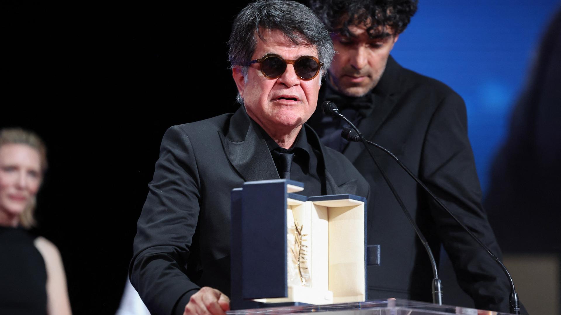 Jafar Panahi steht an einem Rednerpult, er trägt eine Sonnenbrille. Vor ihm steht die Goldene Palme. Jafar Panahi steht an einem Rednerpult, er trägt eine Sonnenbrille. Vor ihm steht die Goldene Palme.