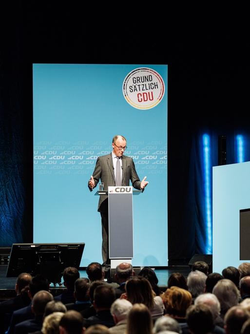 Der CDU-Vorsitzende Friedrich Merz steht vor Publikum an einem Rednerpult, hinter ihm an der Wand ist auf einem runden Schild zu lesen: Grundsätzlich CDU. Rechts am Bühnenrand sind die Farben Schwarz, Rot, Gold als aufstrebende Balkendiagramme zu sehen.