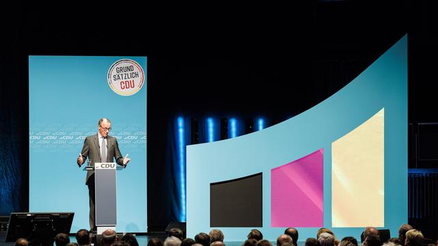 Der CDU-Vorsitzende Friedrich Merz steht vor Publikum an einem Rednerpult, hinter ihm an der Wand ist auf einem runden Schild zu lesen: Grundsätzlich CDU. Rechts am Bühnenrand sind die Farben Schwarz, Rot, Gold als aufstrebende Balkendiagramme zu sehen.