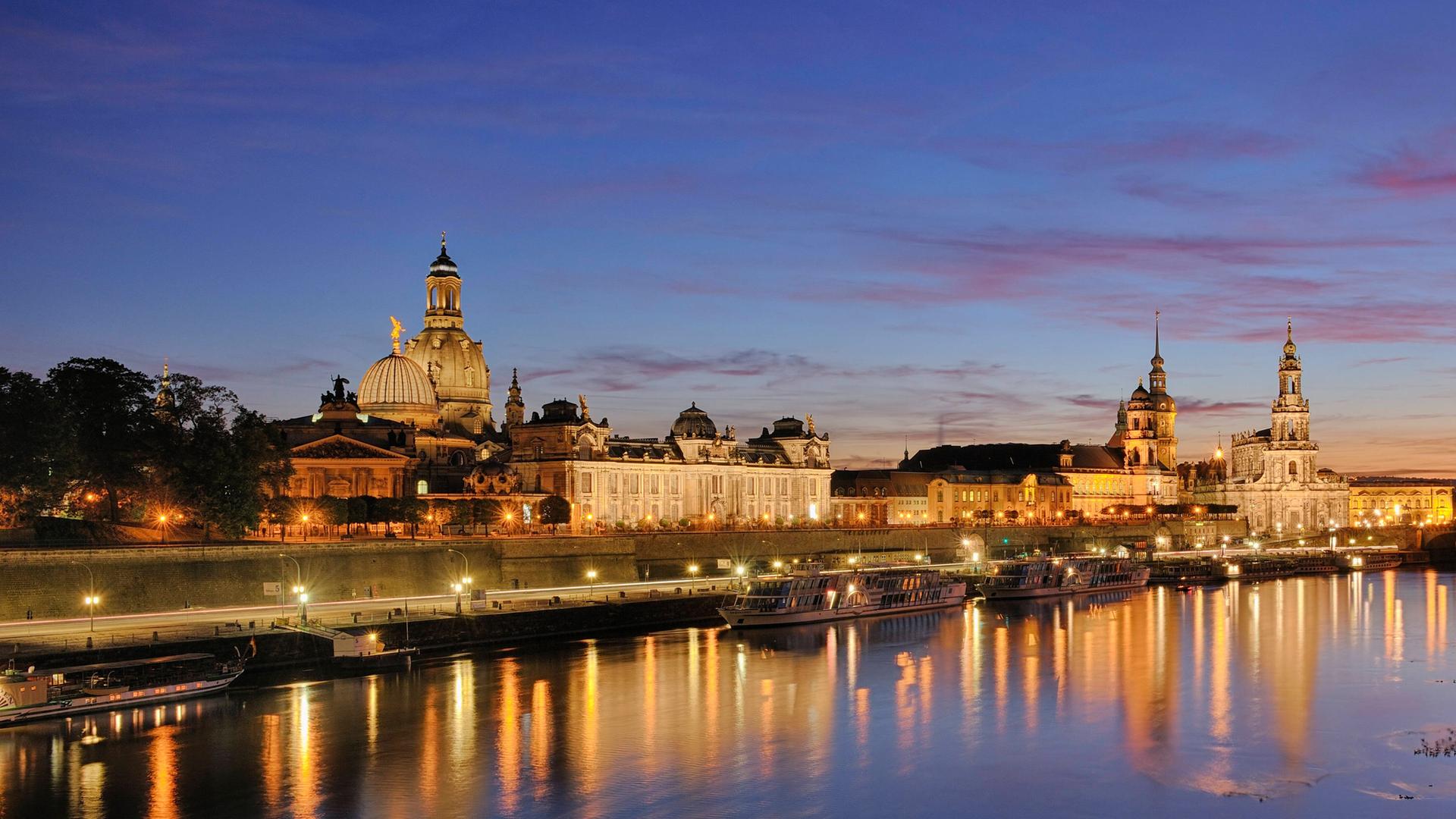 Eine blau-rosa gefärbter Himmel liegt über der Dresdner Stadtkulisse, die man angestrahlt über den Fluß hinweg erblickt.