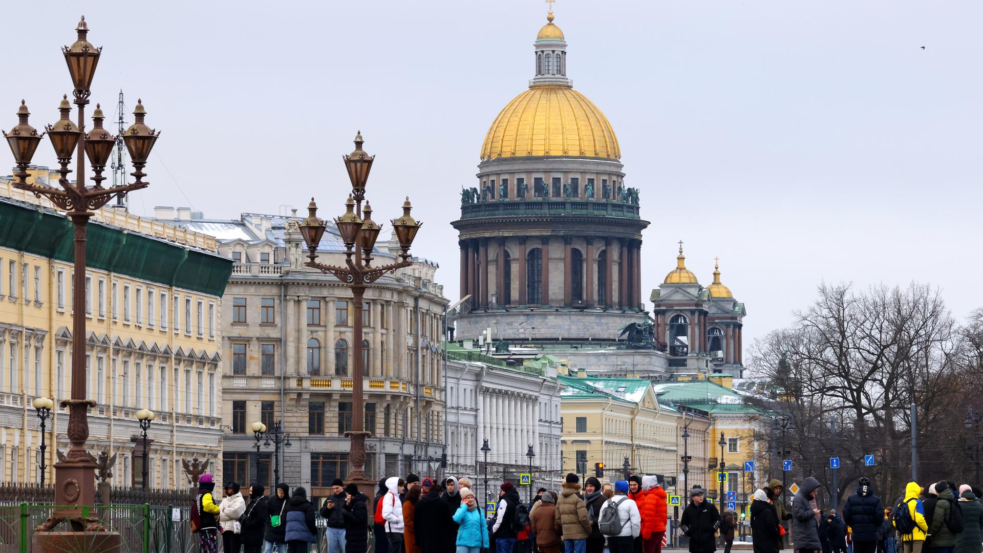 Eine Gruppe von Menschen steht auf einem Platz, im Hintergrund sieht man Häuser, Straßenlaternen und eine Kirche mit einer großen goldenen Kuppel. Eine Gruppe von Menschen steht auf einem Platz, im Hintergrund sieht man Häuser, Straßenlaternen und eine Kirche mit einer großen goldenen Kuppel.
