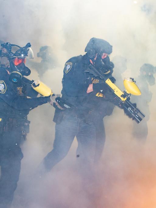 Bei einem Einsatz in Portland, Oregon im Februar 2026 stehen ICE-Agenten in Uniform und mit Masken in einer Wolke aus Tränengas und halten Tränengaswaffen in der Hand.