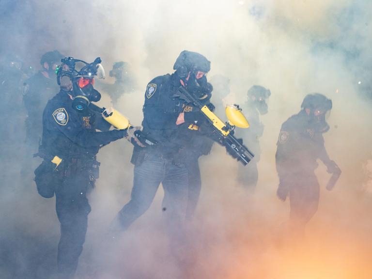 Bei einem Einsatz in Portland, Oregon im Februar 2026 stehen ICE-Agenten in Uniform und mit Masken in einer Wolke aus Tränengas und halten Tränengaswaffen in der Hand.