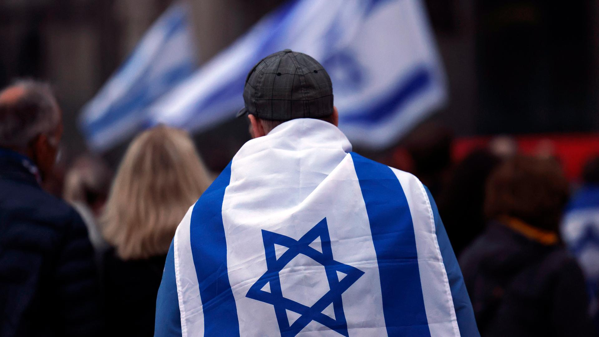 Bei einer Pro-Israel-Veranstaltung trägt ein Mann eine Israel-Flagge um die Schultern. Er ist von hinten zu sehen. Im Hintergrund sind weiter Israel-Flaggen zu sehen. 