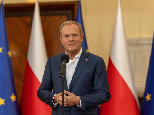 Polens Premier Donald Tusk steht mit gefalteten Händen vor einem Mikrofon.
