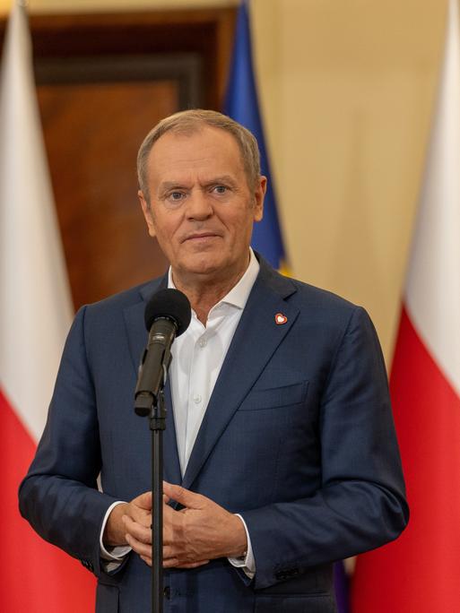 Polens Premier Donald Tusk steht mit gefalteten Händen vor einem Mikrofon.
