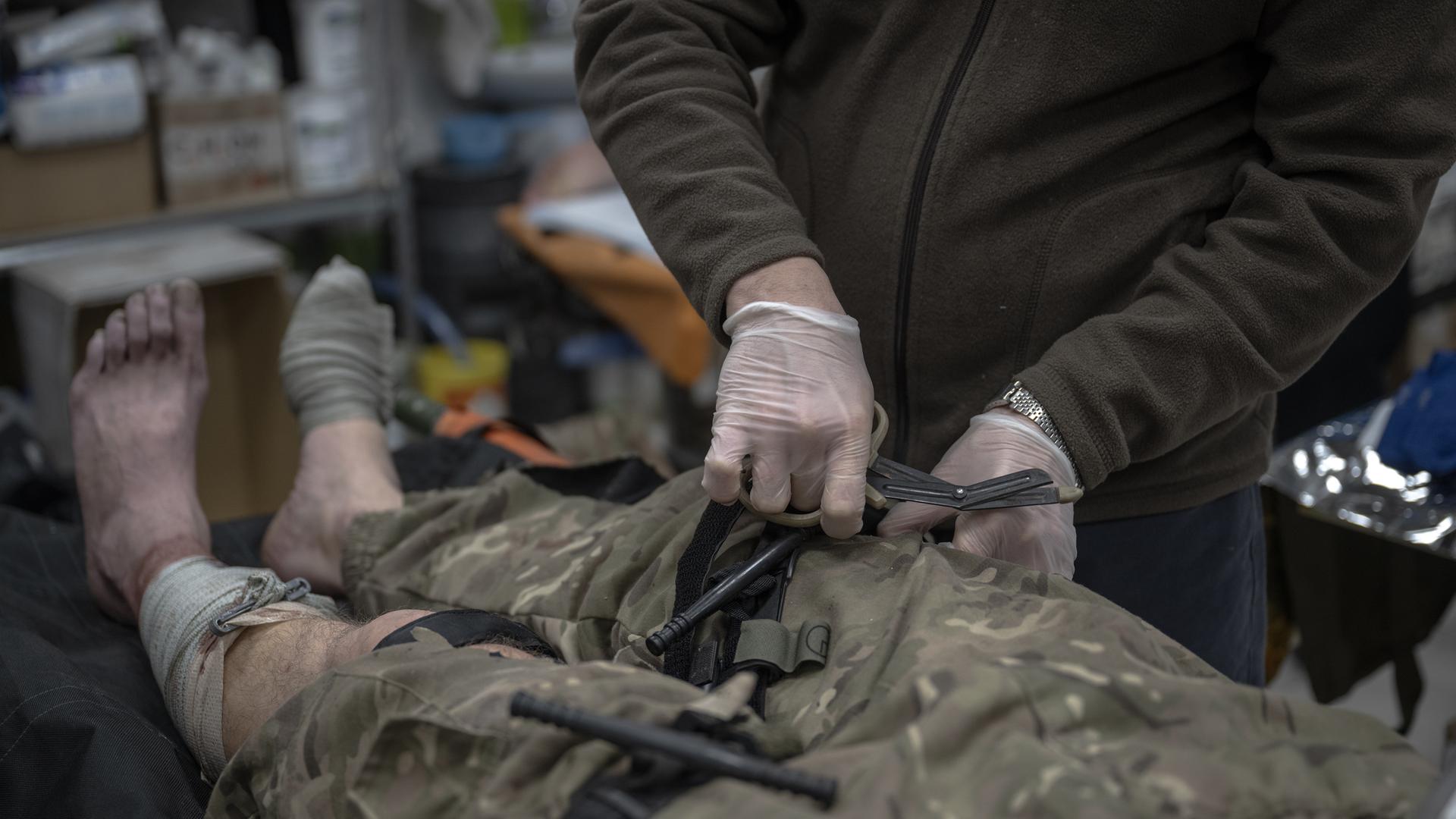 Ein ukrainischer Frontsoldat wird medizinisch behandelt. Man sieht zwei Hände ins Bild hineinreichen, die den Mann am Bein behandeln. Der Soldat auf dem Behandlungstisch hat eine Hose in Tarnfleck an, die Füße sind nackt. Man sieht den Körper hüftabwärts im Bild-