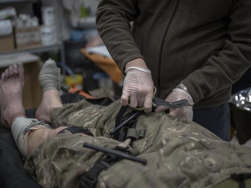 Ein ukrainischer Frontsoldat wird medizinisch behandelt. Man sieht zwei Hände ins Bild hineinreichen, die den Mann am Bein behandeln. Der Soldat auf dem Behandlungstisch hat eine Hose in Tarnfleck an, die Füße sind nackt. Man sieht den Körper hüftabwärts im Bild-