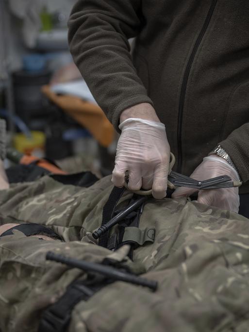 Ein ukrainischer Frontsoldat wird medizinisch behandelt. Man sieht zwei Hände ins Bild hineinreichen, die den Mann am Bein behandeln. Der Soldat auf dem Behandlungstisch hat eine Hose in Tarnfleck an, die Füße sind nackt. Man sieht den Körper hüftabwärts im Bild-