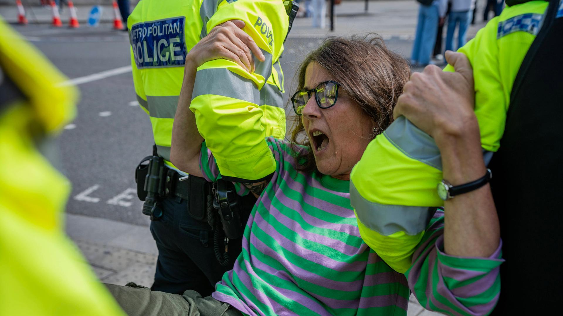 Eine Frau mittleren Alters wird von mehreren Polizisten abtransportiert. Sie trägt einen gestreiften Pulli, eine Jeans und eine Brille und schreit offenbar.
