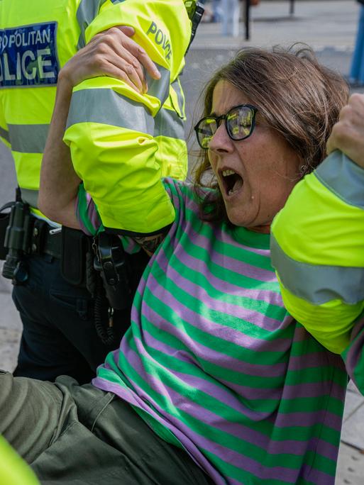 Eine Frau mittleren Alters wird von mehreren Polizisten abtransportiert. Sie trägt einen gestreiften Pulli, eine Jeans und eine Brille und schreit offenbar.