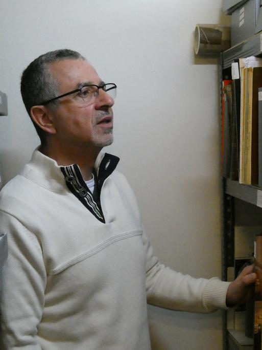 Ein Mann mit kurzem, ergrauten Haar und Brille vor einem Archiv-Regal