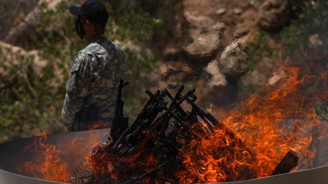 In einer Feuerschale brennen Waffen. Ein Soldat steht hinter der Feuerschale. 