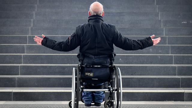 Ein Mann im Roll-Stuhl steht vor einer Treppe. Er kommt nicht weiter. Er streckt verzweifelt die Arme aus. 