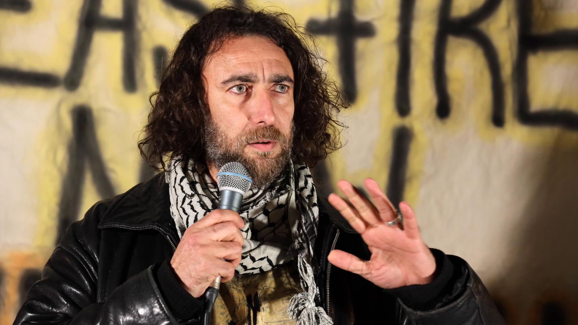 Ghayath Almadhoun mit lockigem schulterlangen Haar und Palästinesertuch spricht bei einer Kundgebung vor einem Banner in eine Mikrofon Ghayath Almadhoun mit lockigem schulterlangen Haar und Palästinesertuch spricht bei einer Kundgebung vor einem Banner in eine Mikrofon