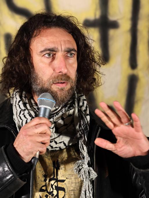 Ghayath Almadhoun mit lockigem schulterlangen Haar und Palästinesertuch spricht bei einer Kundgebung vor einem Banner in eine Mikrofon