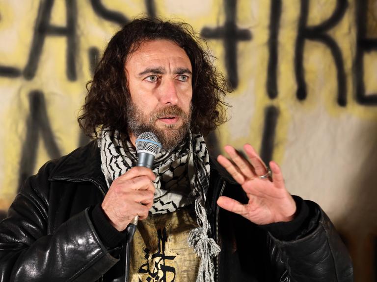 Ghayath Almadhoun mit lockigem schulterlangen Haar und Palästinesertuch spricht bei einer Kundgebung vor einem Banner in eine Mikrofon