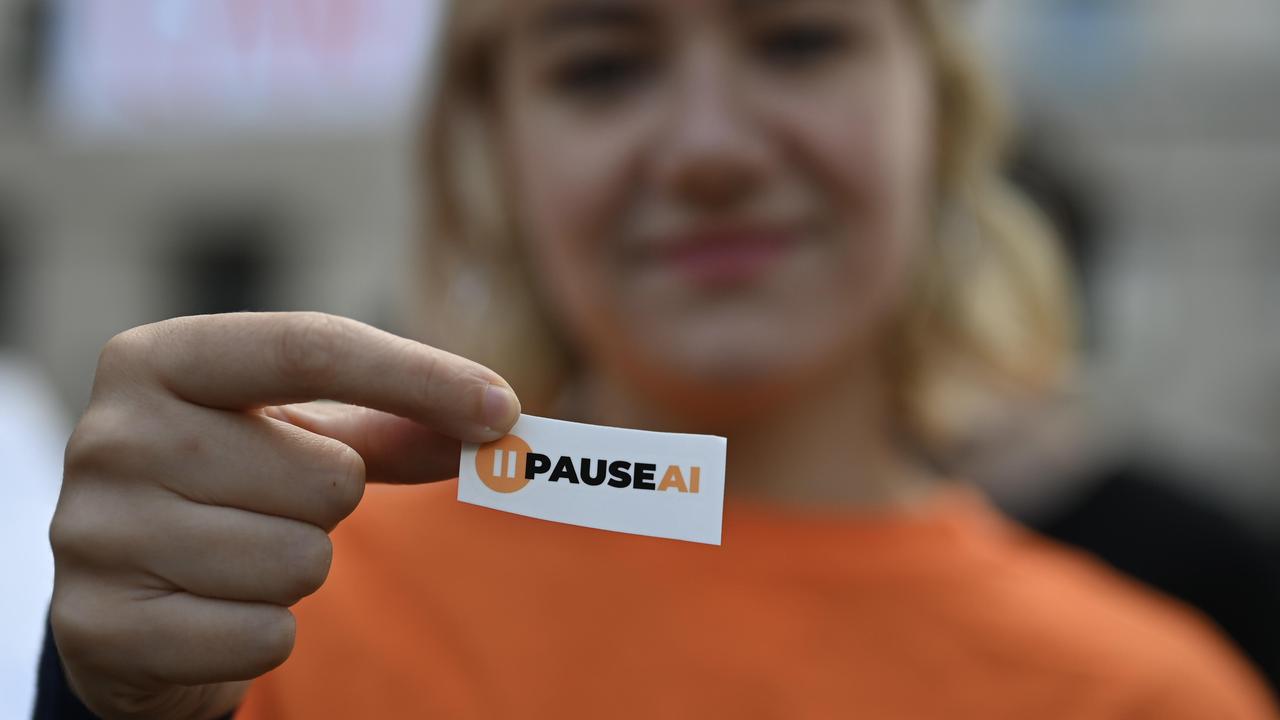 Eine KI-Sicherheit-Aktivistin hält bei einer Demonstration in der britischen Hauptstadt London ein kleines Schild in die Kamera, auf dem "PAUSEAI" steht. In der Unschärfe ist im Hintergrund ihr Gesicht zu sehen.