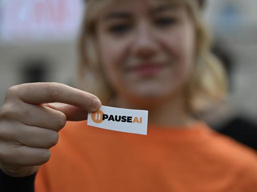 Eine KI-Sicherheit-Aktivistin hält bei einer Demonstration in der britischen Hauptstadt London ein kleines Schild in die Kamera, auf dem "PAUSEAI" steht. In der Unschärfe ist im Hintergrund ihr Gesicht zu sehen. Eine KI-Sicherheit-Aktivistin hält bei einer Demonstration in der britischen Hauptstadt London ein kleines Schild in die Kamera, auf dem "PAUSEAI" steht. In der Unschärfe ist im Hintergrund ihr Gesicht zu sehen.