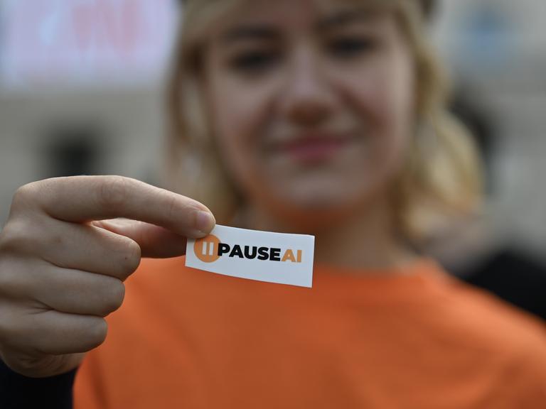 Eine KI-Sicherheit-Aktivistin hält bei einer Demonstration in der britischen Hauptstadt London ein kleines Schild in die Kamera, auf dem "PAUSEAI" steht. In der Unschärfe ist im Hintergrund ihr Gesicht zu sehen.