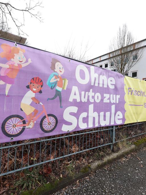 Vor einer Schule hängt ein Banner an einem Zaun. Darauf steht: Ohne Auto zur Schule. Frische Luft statt Elterntaxi"
