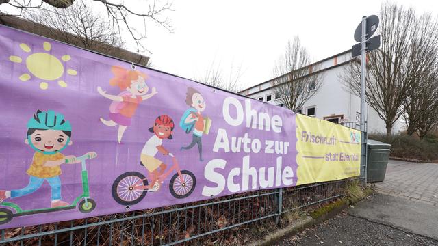 Vor einer Schule hängt ein Banner an einem Zaun. Darauf steht: Ohne Auto zur Schule. Frische Luft statt Elterntaxi"