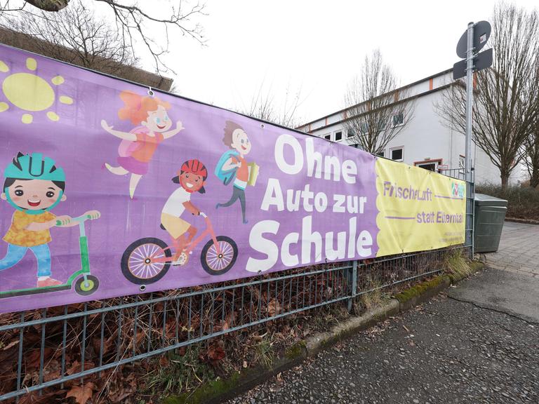 Vor einer Schule hängt ein Banner an einem Zaun. Darauf steht: Ohne Auto zur Schule. Frische Luft statt Elterntaxi"