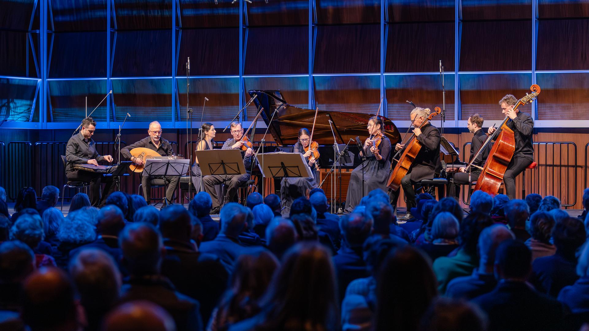 Ein Kanun-Spieler, ein Oud-Musiker, ein Klavierduo, ein Streichquartett und ein Kontrabassist musizieren auf einer blau erleuchteten Bühne vor Publikum Ein Kanun-Spieler, ein Oud-Musiker, ein Klavierduo, ein Streichquartett und ein Kontrabassist musizieren auf einer blau erleuchteten Bühne vor Publikum